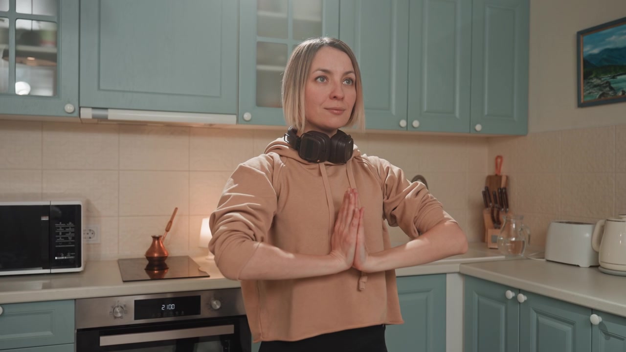 Donna in cucina che medita con la musica, cuffie appoggiate al collo, luce soffusa della lampada, mobili color verde acqua, lenti gesti delle mani simili a una preghiera, calma introspettiva e atmosfera domestica accogliente.