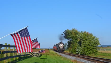 Ein-Blick-Auf-Eine-Reihe-Sanft-Wehender-Amerikanischer-Flaggen-Auf-Einem-Zaun-Neben-Ackerland,-Während-Sich-Während-Der-Goldenen-Stunde-Ein-Dampf-Personenzug-Nähert,-Der-Rauch-Bläst