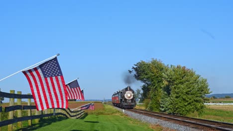 a view of a line of gently waving american flags on a fence by farmlands as a steam passenger train blowing smoke approaches during the golden hour