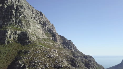 Aerial-drone-flight-along-Table-Mountain-rocks-and-epic-reveal-of-Lion's-Head-in-far-background-against-Atlantic-Ocean-panorama-and-bright-blue-sky