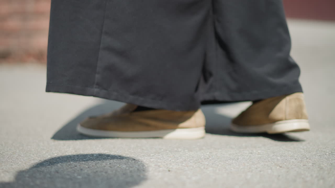 Close up of person legs moving slightly on sunlit pavement, wearing black pants and brown shoes, subtle shifting shows nervous energy, curiosity, and quiet emotion through light