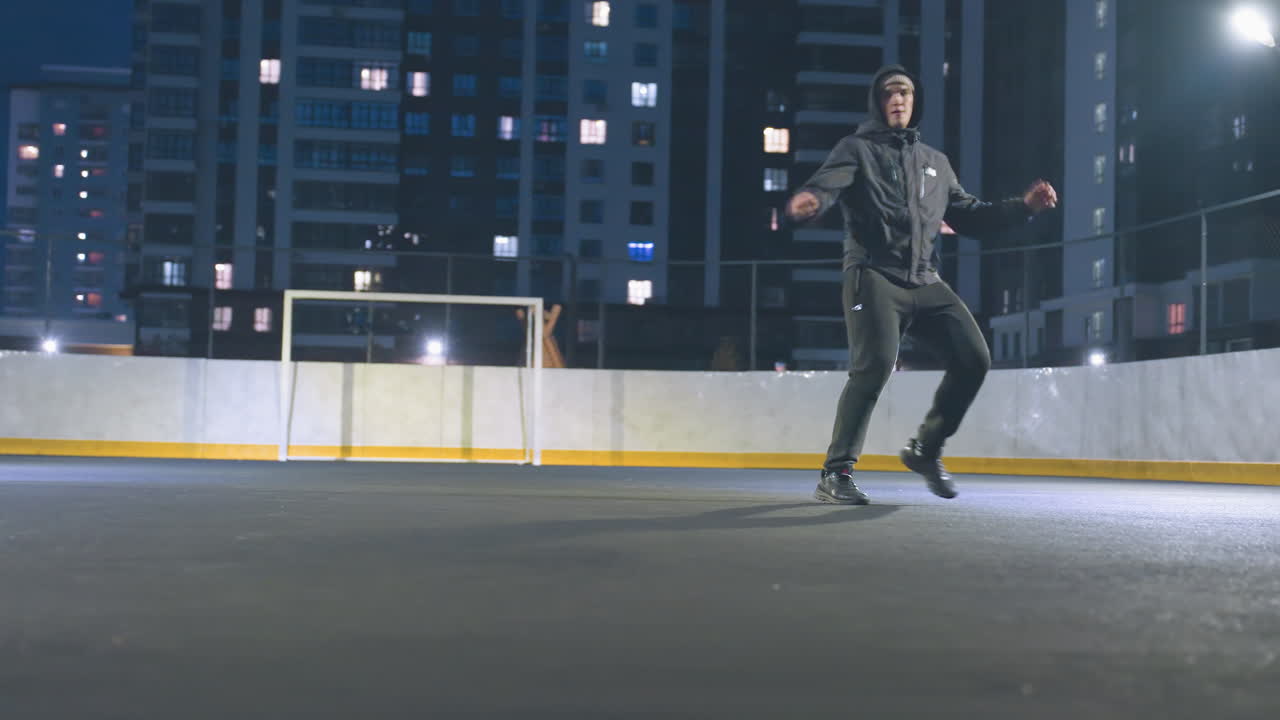 Man in hoodie kicks a soccer ball, with background featuring goal post behind and street light, surrounded by illuminated field and cityscape