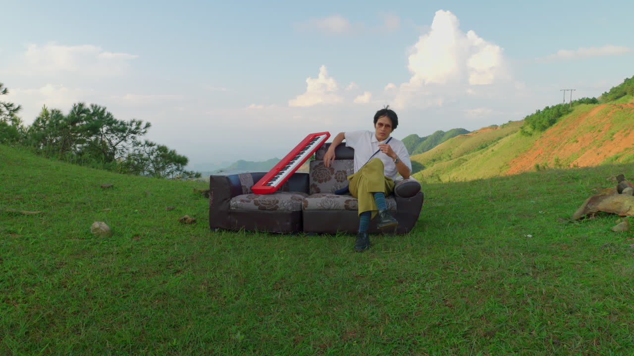 young asiatic artist musician looking for inspiration while sitting on couch in outdoor mountains valley during a day of sun with a keyboard piano Musical instrument