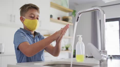 Caucasian-boy-wearing-yellow-facemask-standing-at-kitchen-sink-washing-hands-under-running-tap