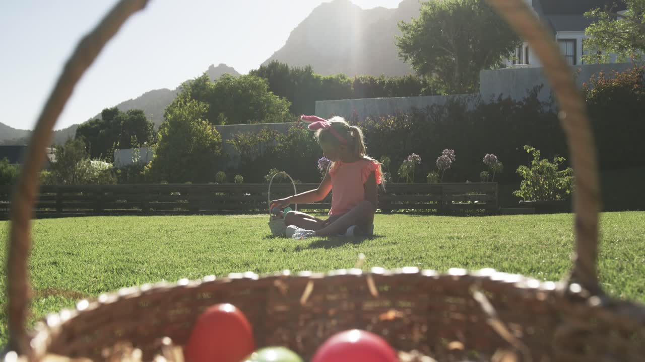 Spotting colorful eggs, child sitting on lawn reaching into wire basket, inspecting egg for Easter