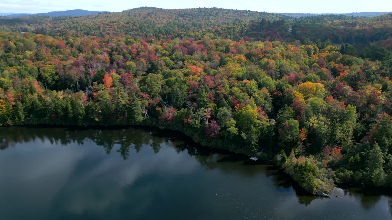 Aerial pan across lake edge with early fall colors reflecting sky and clouds on clear water, New England mountains