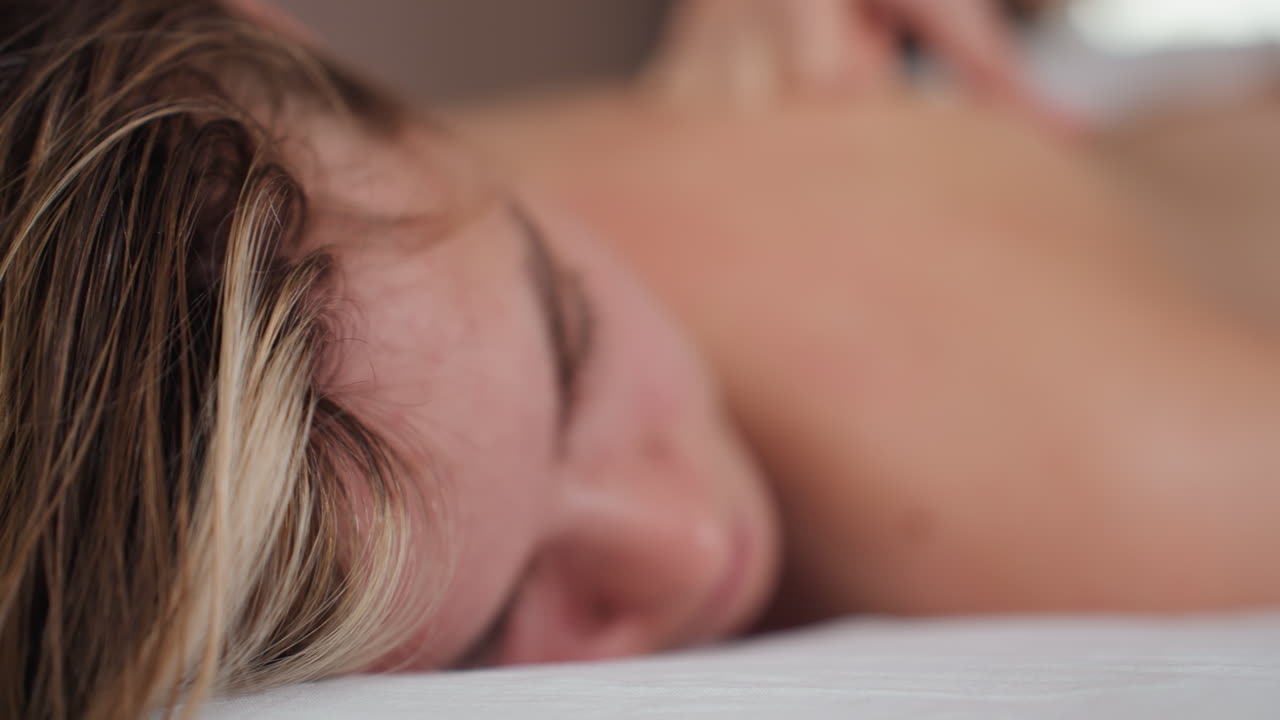 Closeup view of blond woman lying face down with closed eyes, relaxed expression, and blurred background creating soft focus on facial features during calm, quiet moment on massage table