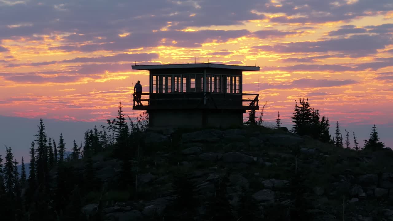 Un uomo cammina sulla veranda del balcone di una torre di avvistamento antincendio isolata sulla cima di una montagna nel remoto nord dell'Idaho e si appoggia alla ringhiera di fronte a una spettacolare alba o tramonto dorato e colorato