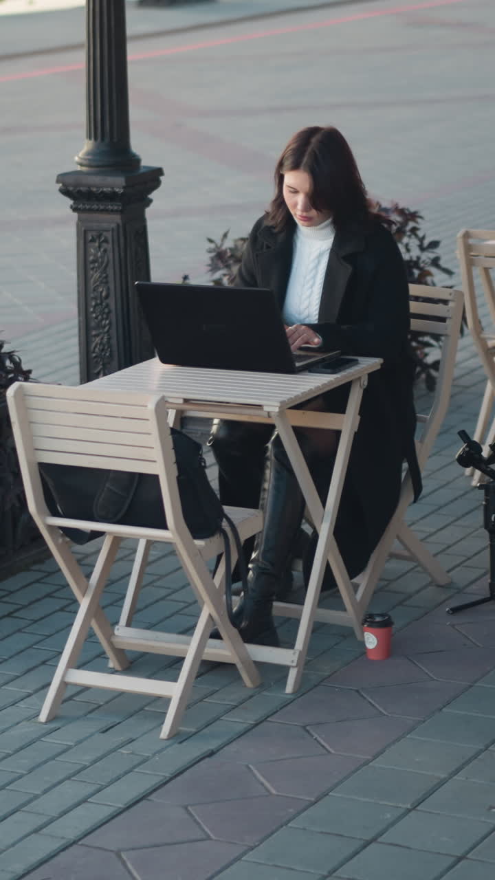 Young woman working on laptop outdoors at a restaurant surrounded by wooden chairs, plants, and a tripod, she is seated at a table with a coffee cup, with a paved walkway and blurred background