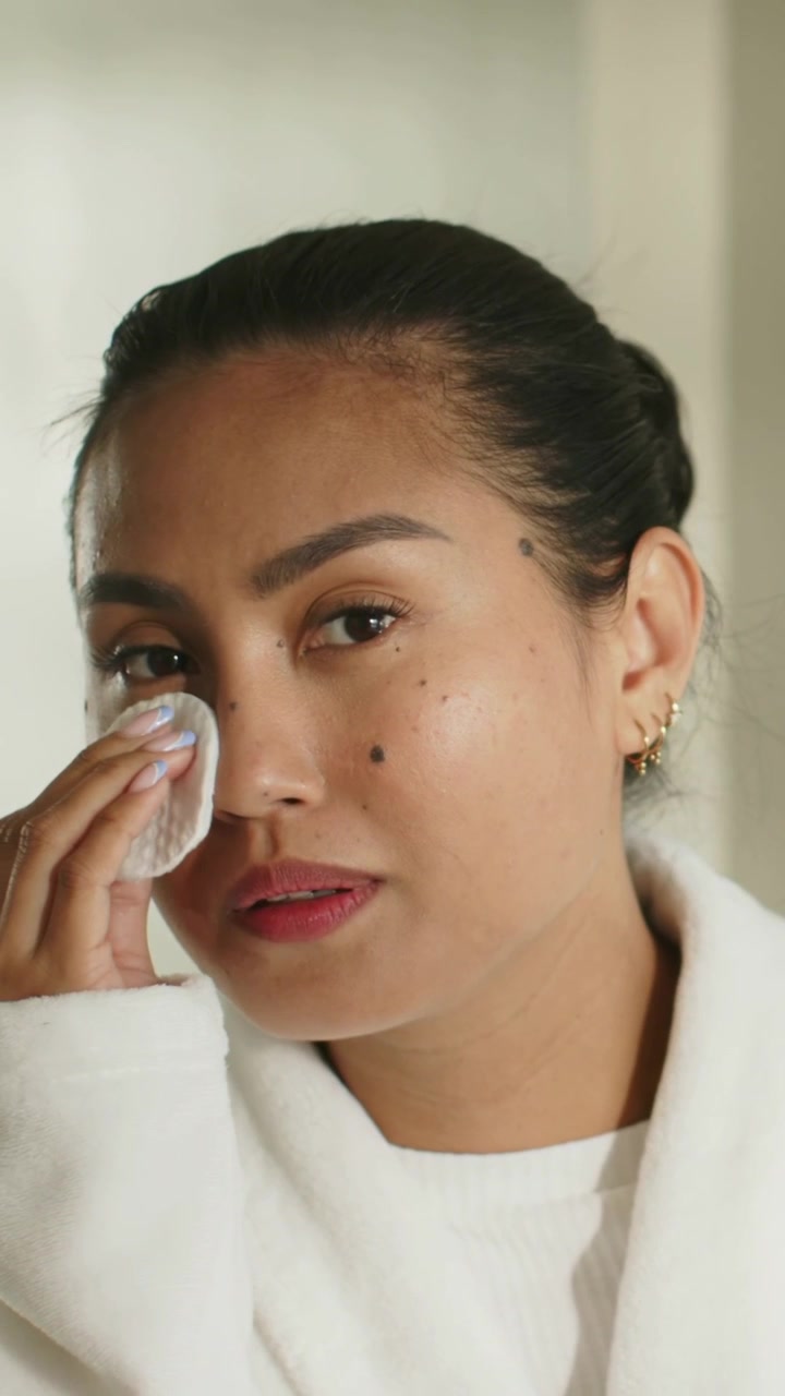 Vertical video: Starting mid adult Asian woman removing makeup in robe at home, showing cotton pads