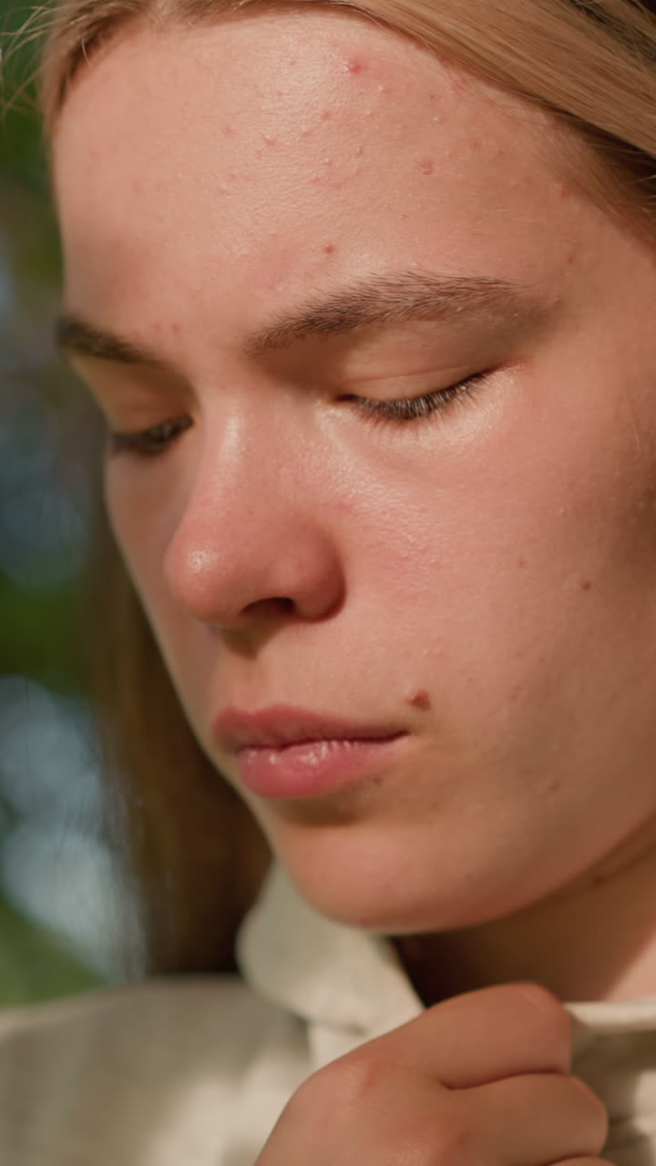 은 머리카락을 가진 젊은 여성의 클로즈업 깊은 생각에, 부드럽게 그녀의 후드 목걸이를 만지, 나무 잎을 통해 필터링 빛으로 부드럽게 조명 된 얼굴