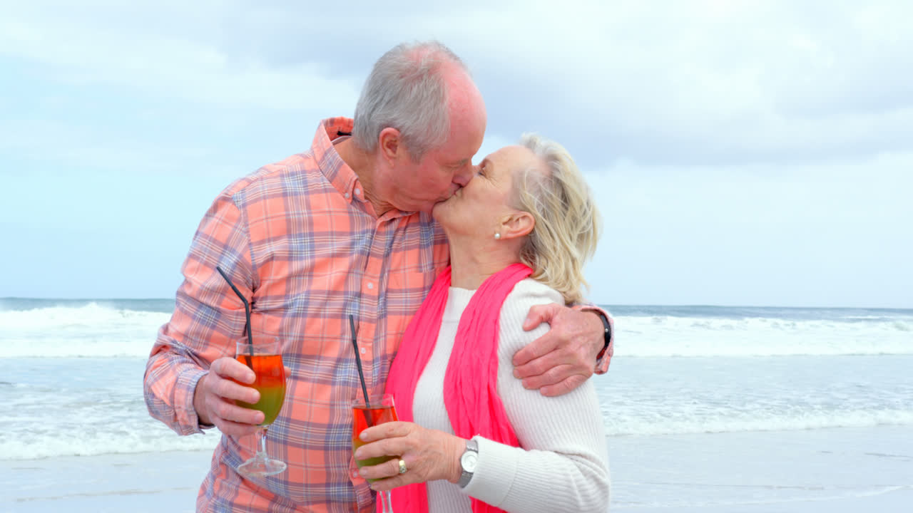 Front view of old caucasian senior couple kissing each other and drinking cocktail at beach 4k