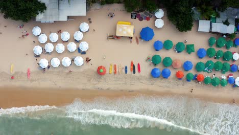 Hermosa-Vista-Aérea-De-Drones-En-Ascenso-Vista-Superior-De-Kayaks-Coloridos-Para-Alquilar-Sentados-En-La-Arena-En-La-Playa-Tropical-Praia-Do-Madeiro-Con-Sombrillas-Coloridas-Y-Agua-Azul-Clara-Cerca-De-Pipa-En-Brasil