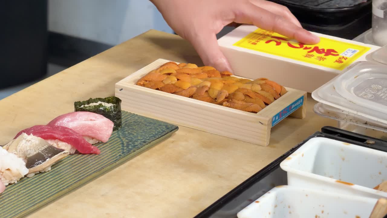 Close-up of chef opening wooden sea urchin box beside assorted sushi under bright kitchen lighting
