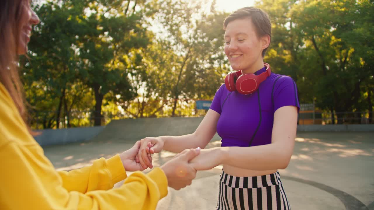 disparando una vista de cerca de una chica feliz en una camiseta púrpura y auriculares rojos que la ayudan a patinar una chica morena en un suéter blanco en un parque de skate en verano