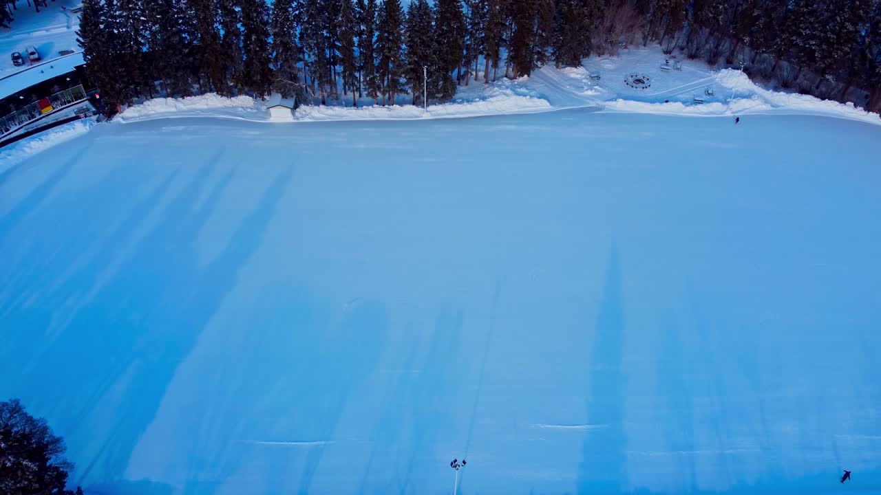 Aerial winter rise over massive outdoor skating rink in a snow covered city park during sunset shades of shadows on ice surrounded by tall pine fir trees and hardly any people outside panoramic view