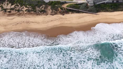 Drone camera glides above cliffside buildings, sandy beach, and approaching ocean waves in daylight