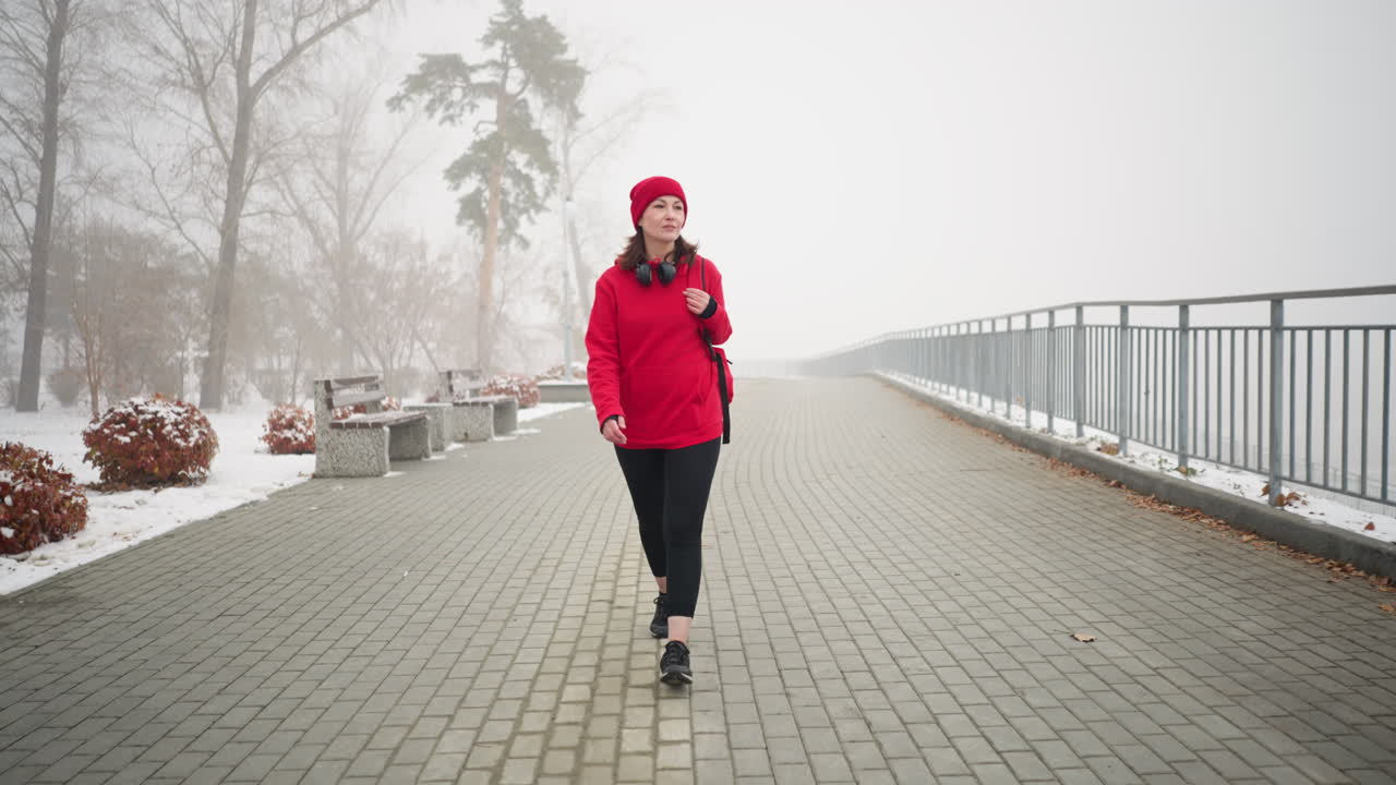 mujer con chaqueta roja de invierno y calentador de cabeza con auriculares en el cuello, caminando por un camino cubierto de nieve y niebla, bancos y árboles llenos de nieve crean una atmósfera serena