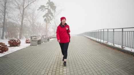 woman wearing red winter jacket and head warmer with headphones on her neck, walking along snowy fog-covered path, benches and trees dusted with snow create serene atmosphere