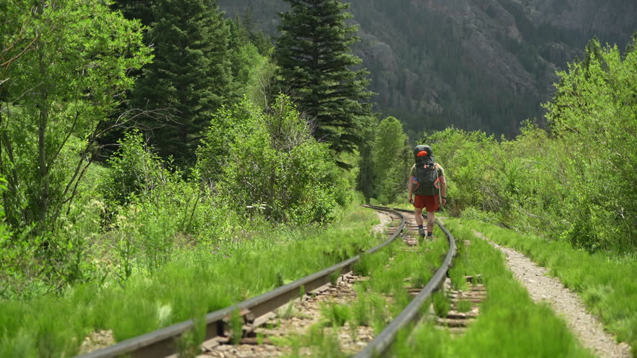 vista trasera de un excursionista con mochila caminando por un antiguo ferrocarril industrial en un paisaje verde