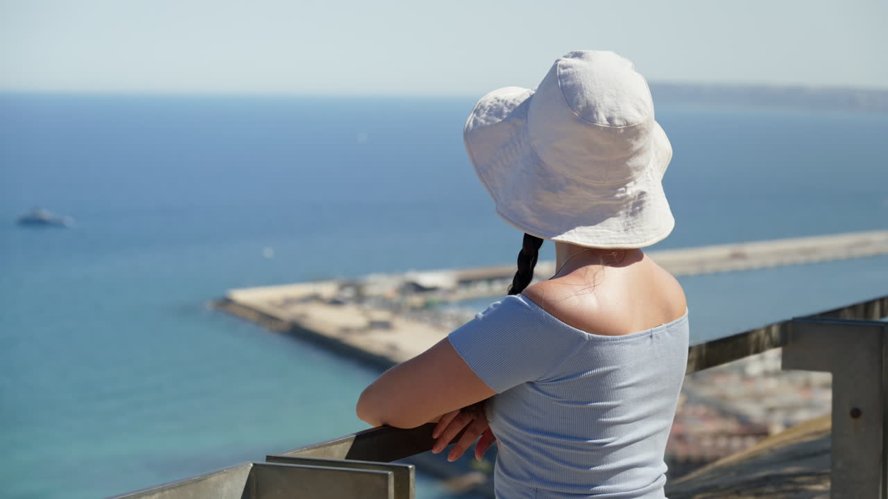 A traveler girl in a white bucket hat takes in the coast from a breezy hilltop lookout in Alicante, Spain