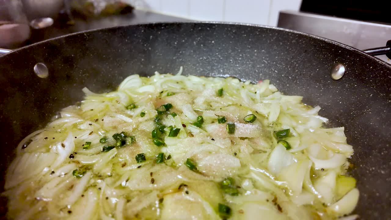 Cebolas fatiadas e pimentas verdes fritando em óleo quente, criando vapor em uma panela