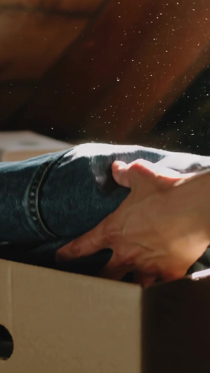 Vertical video: Placing hands lifting jeans into box in loft, smoothing near books for donation