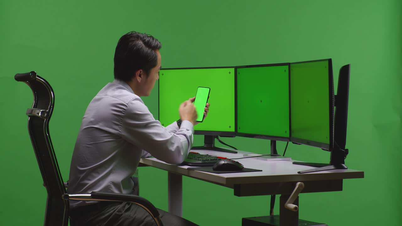 hombre de negocios revisando un teléfono inteligente en una estación de trabajo de computadora con múltiples monitores en un estudio de pantalla verde