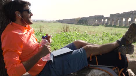 cyclist with bike sitting on a rock under a tree listening to music with his laptop and drinking water in front of ancient roman aqueduct. young attractive athletic man with orange sportswear headphones and backpack in parco degli acquedotti in rome