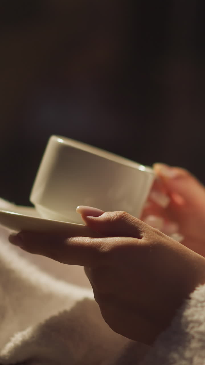 Resting woman in bathrobe drinks hot beverage in cup enjoying being alone. Calm and quiet atmosphere in semi lit hotel room. Peace of mind alone with thoughts