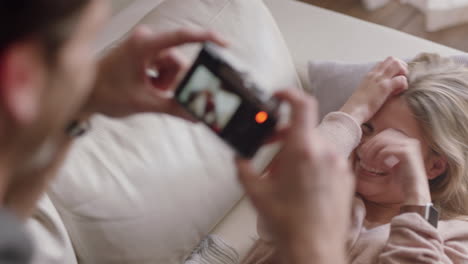 happy young couple taking photos together using camera having fun at home on sofa playfully enjoying romantic relationship photographing each other making faces