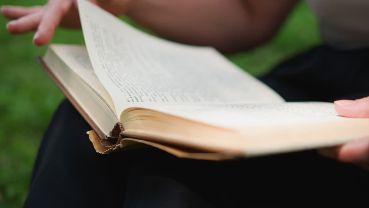 Close up partial view of lady flipping through old book pages while sitting outdoors on green grass, sunlight highlighting motion of gentle hands turning pages