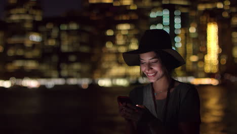 Frau-Benutzt-Nachts-Ihr-Mobiltelefon-Mit-Der-Skyline-Der-Stadt-Im-Hintergrund