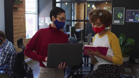 Diverse-female-colleagues-wearing-face-masks-using-laptop-taking-notes-discussing-at-modern-office