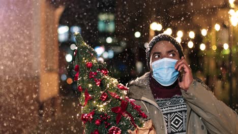 portrait of young african american man wearing facial mask talking on smartphone and holding a christmas tree on the street while it¬¥s snowing in christmas