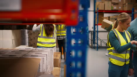 Team-Leader-With-Digital-Tablet-In-Busy-Warehouse-Training-Female-Intern-Standing-By-Shelves