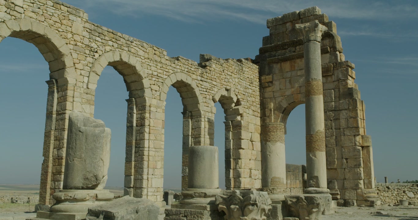 Elokuvamainen näkymä Volubilisin basilikasta, esitellen monumentaalisia roomalaisia kaaria, pylväitä ja säilynyttä kivirakennustaidetta Marokon UNESCOn maailmanperintökohteessa, arkeologisella alueella.