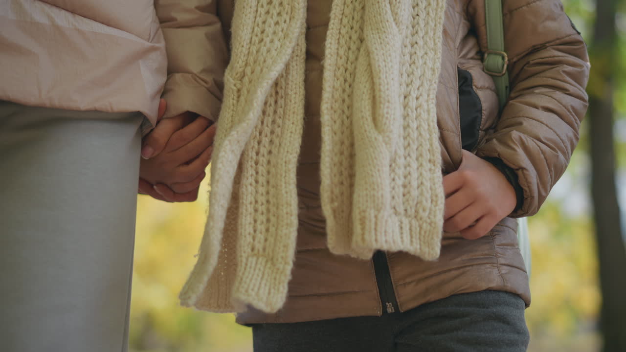 close up of adult and child holding hands walking through autumn park with dry leaves on ground and blurred yellow foliage background wearing warm jackets and knit scarf