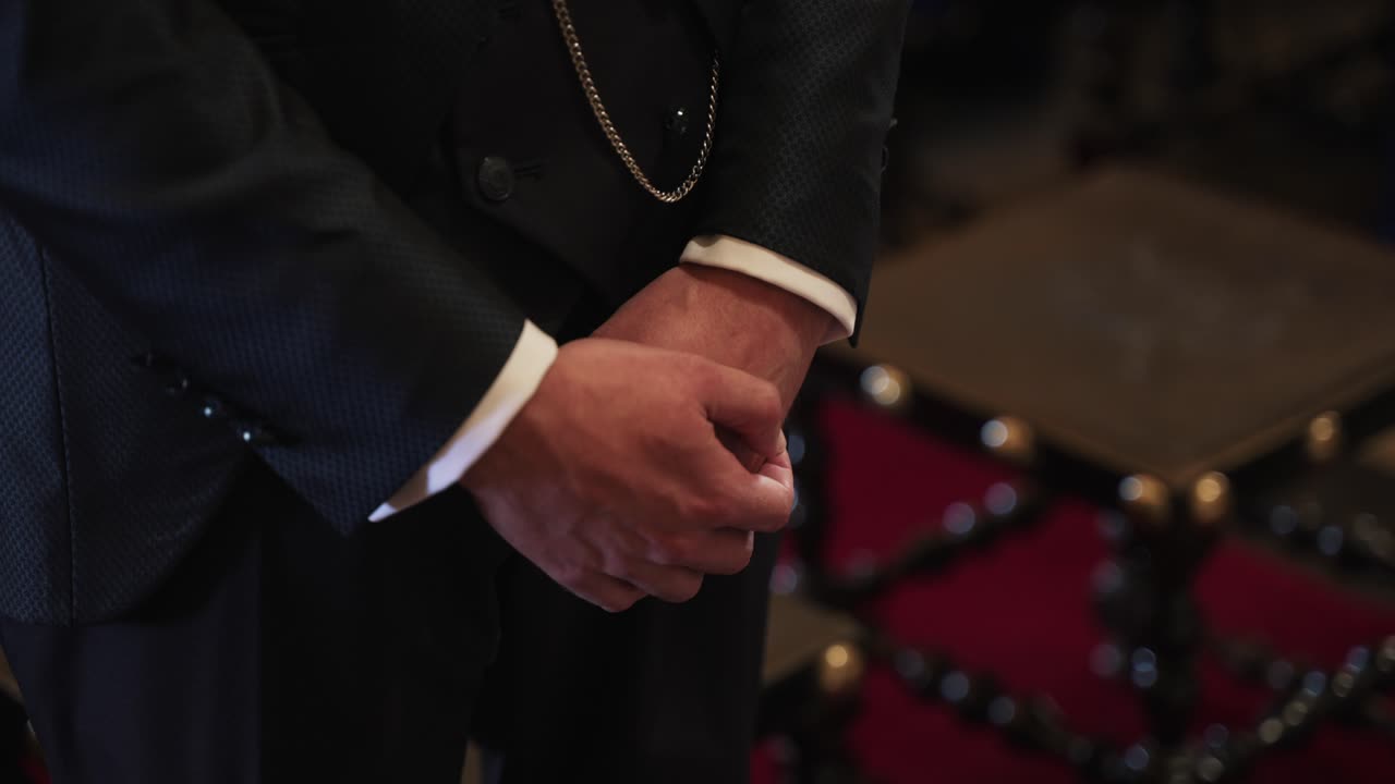 Groom in elegant suit clasps hands while waiting at ceremony aisle