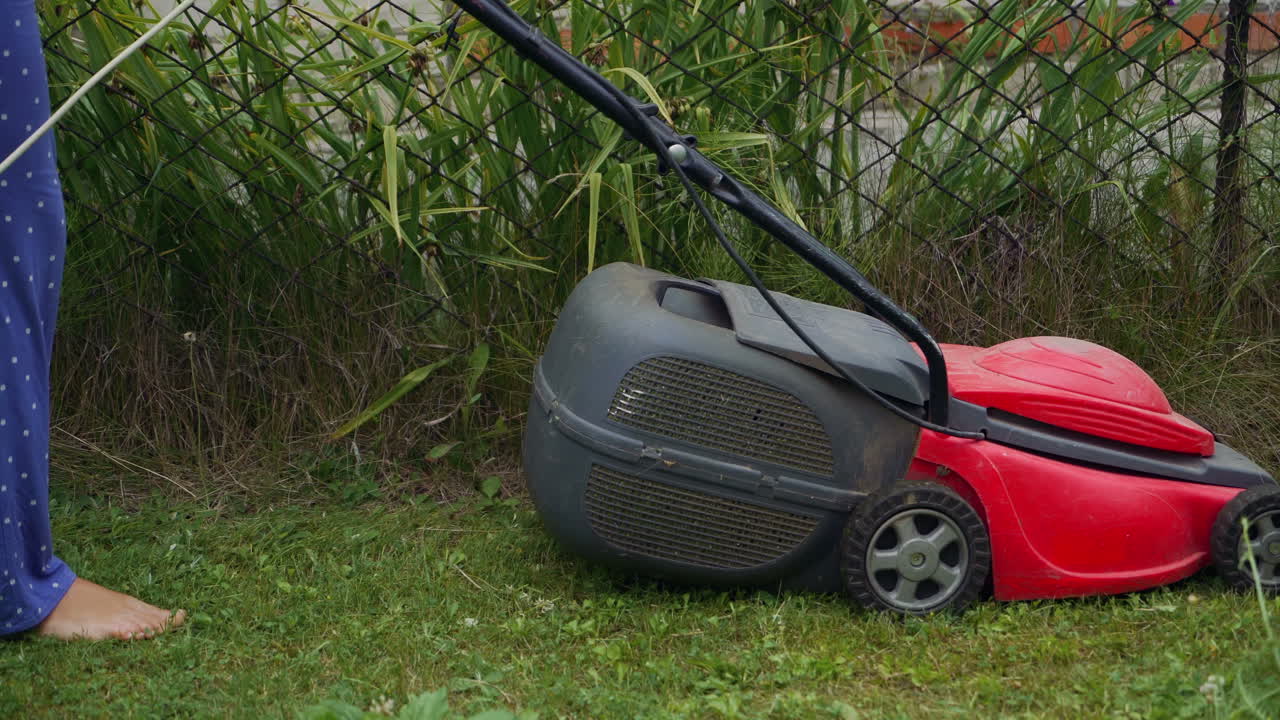 Barefoot young girl mowing grass with a lawn mower in garden. Bare female feet walking behind lawn mower in back yard