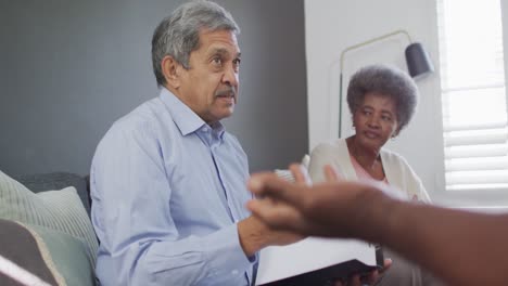 Mixed-race-senior-male-counsellor-with-clipboard-advising-to-group-of-diverse-senior-friends