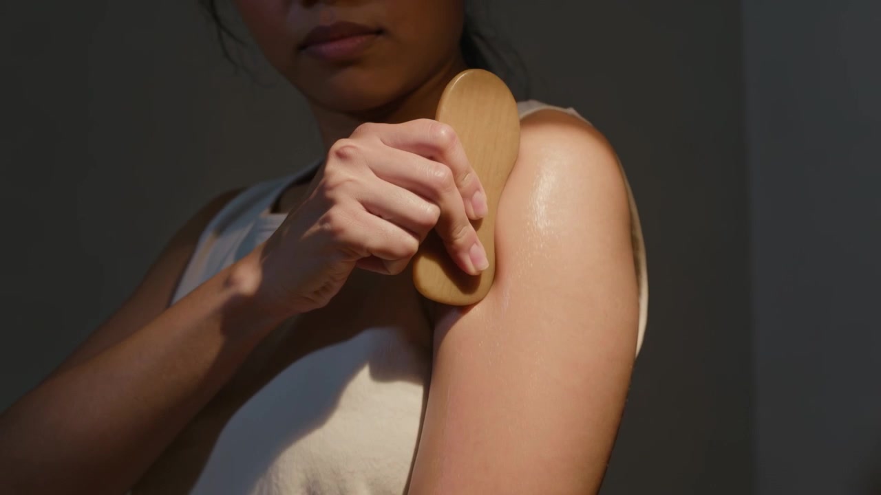 Donna con top senza maniche che preme uno strumento ovale di legno, auto-raschiandosi il braccio con olio in una stanza poco illuminata.
