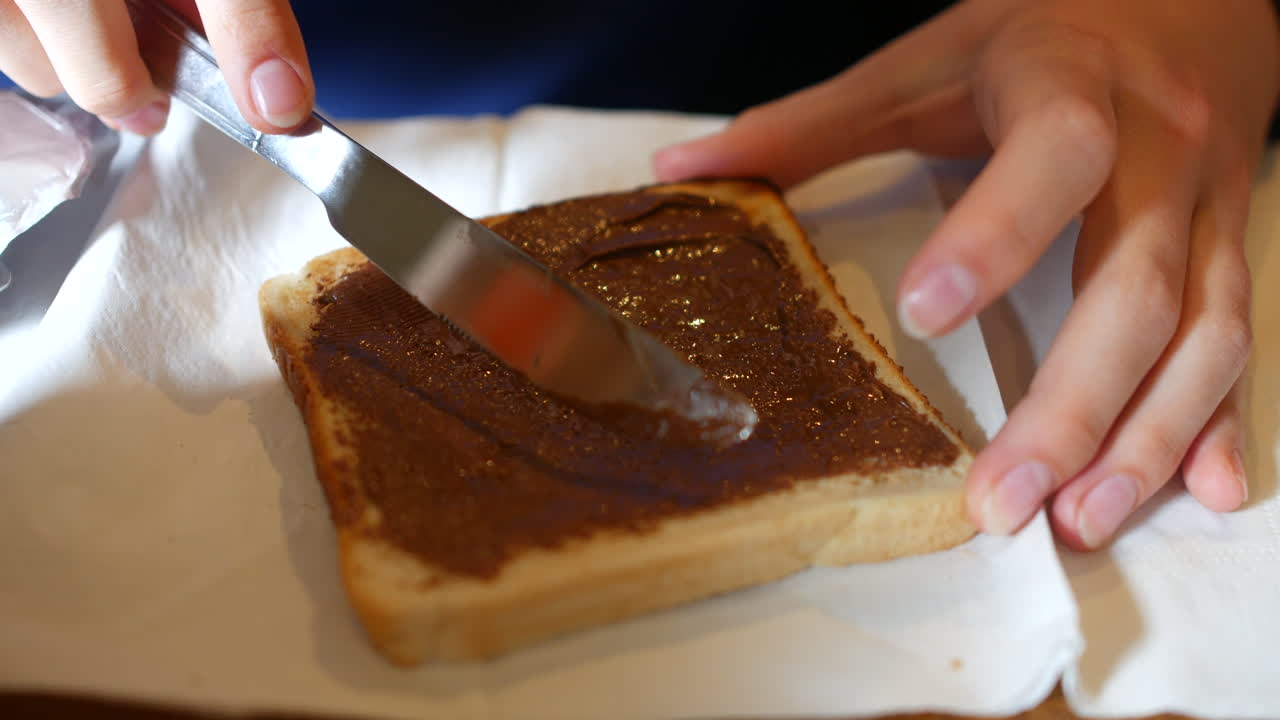 A slice of white bread being spread with chocolate hazelnut spread using a knife, with hands holding the toast on a napkin-covered table surface