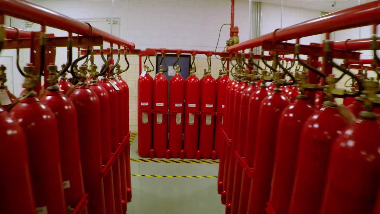 Fileiras de cilindros de gás vermelhos conectados a um sistema de tubulação suspenso, mostrando válvulas, conexões de pressão e armazenamento organizado de cilindros dentro de uma instalação industrial
