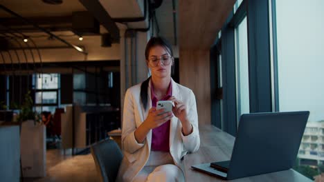 Porträt-Eines-Brünetten-Mädchens-Mit-Runder-Brille,-Weißer-Jacke-Und-Rosa-Hemd,-Das-In-Einem-Weißen-Telefon-Neben-Einem-Panoramafenster-In-Einem-Modernen-Büro-Arbeitet