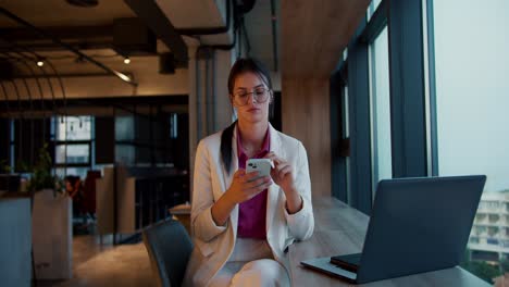 retrato de una chica morena con gafas redondas en una chaqueta blanca y una camisa rosa que trabaja en un teléfono blanco cerca de una ventana panorámica en una oficina moderna