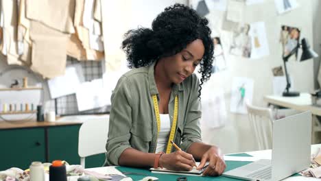 Mujer-Sastre-Sentada-En-La-Mesa-Tomando-Notas-En-El-Taller-De-Costura-Y-Sonríe-A-La-Cámara