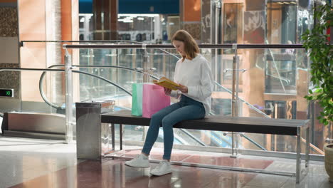 young woman seated in modern shopping mall reading yellow book while swinging her leg slightly, surrounded by colorful shopping bags, glass railings, potted plant, escalator