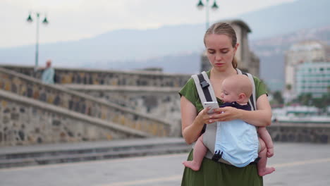 Eine-Junge-Frau-Trägt-Ihr-Kleinkind-In-Einem-Känguru-Rucksack-Und-Fotografiert-Ihre-Reise-Mit-Einem-Mobiltelefon.-Sie-Geht-Und-Schaut-Gelegentlich-Auf-Den-Telefonbildschirm