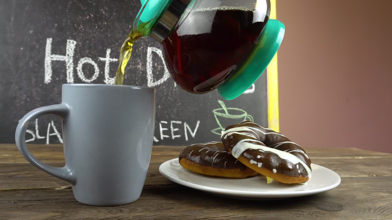 una taza de té y rosquillas en glaseado de chocolate en una mesa de café. el té está derramando de una tetera transparente.